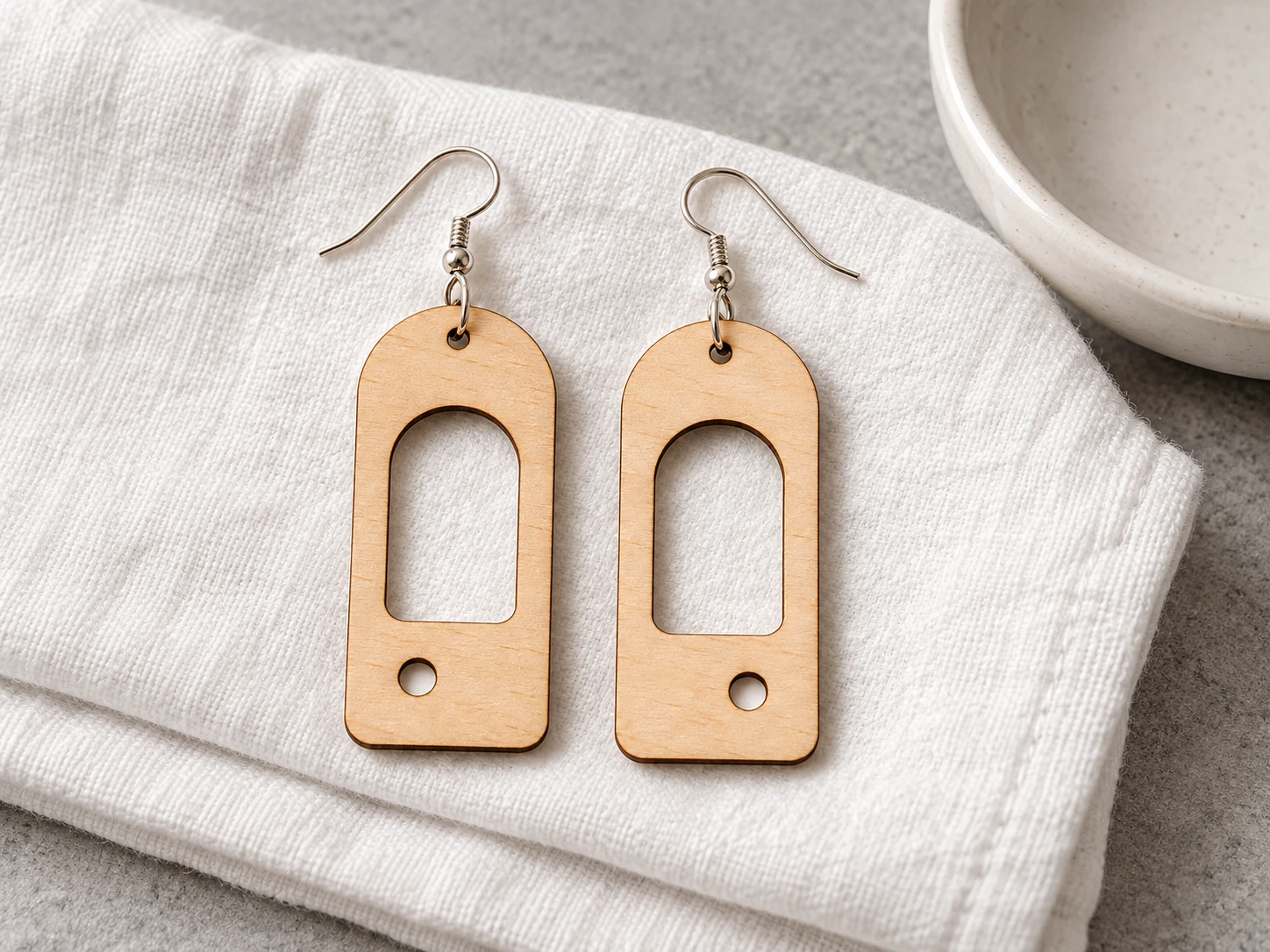 Arc and Orbit wooden arch earrings with circular cutouts resting on ivory linen beside a white jewellery dish.